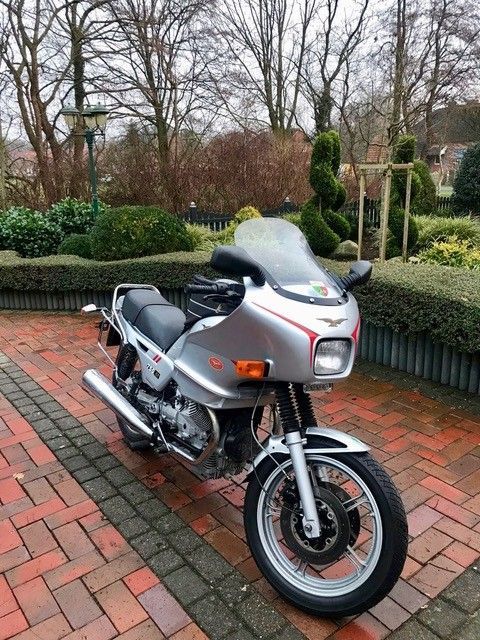 Ein silbernes Tourenmotorrad parkte auf einem roten Backsteinweg in einem angelegten Garten, dahinter befanden sich kahle Bäume und Hecken.