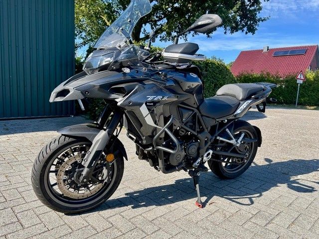 Ein schwarzes Adventure-Motorrad parkt auf einer sonnigen Auffahrt neben einem blauen Gebäude und einem Haus mit rotem Dach.