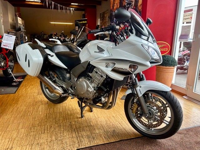 Ein silbernes Honda-Tourenmotorrad steht in einem Innenraum auf beigem Boden, dahinter befinden sich weiße Seitenkoffer und rote Wände.