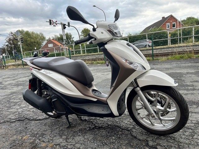 Ein weißer Roller steht auf einem rissigen Gehweg neben einer Straße; im Hintergrund sind Häuser und Bäume zu sehen.