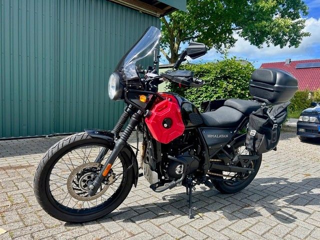 Schwarzes Adventure-Motorrad mit roter Seitentasche parkt auf einer gepflasterten Auffahrt neben einem grünen Gebäude.