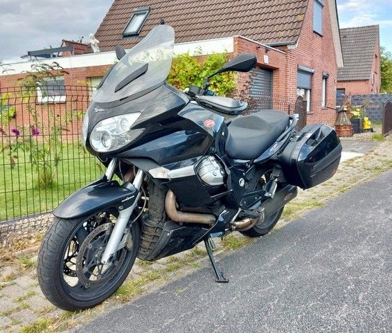 Schwarzes Moto Guzzi-Motorrad, geparkt auf einer asphaltierten Straße vor einem roten Backsteingebäude.