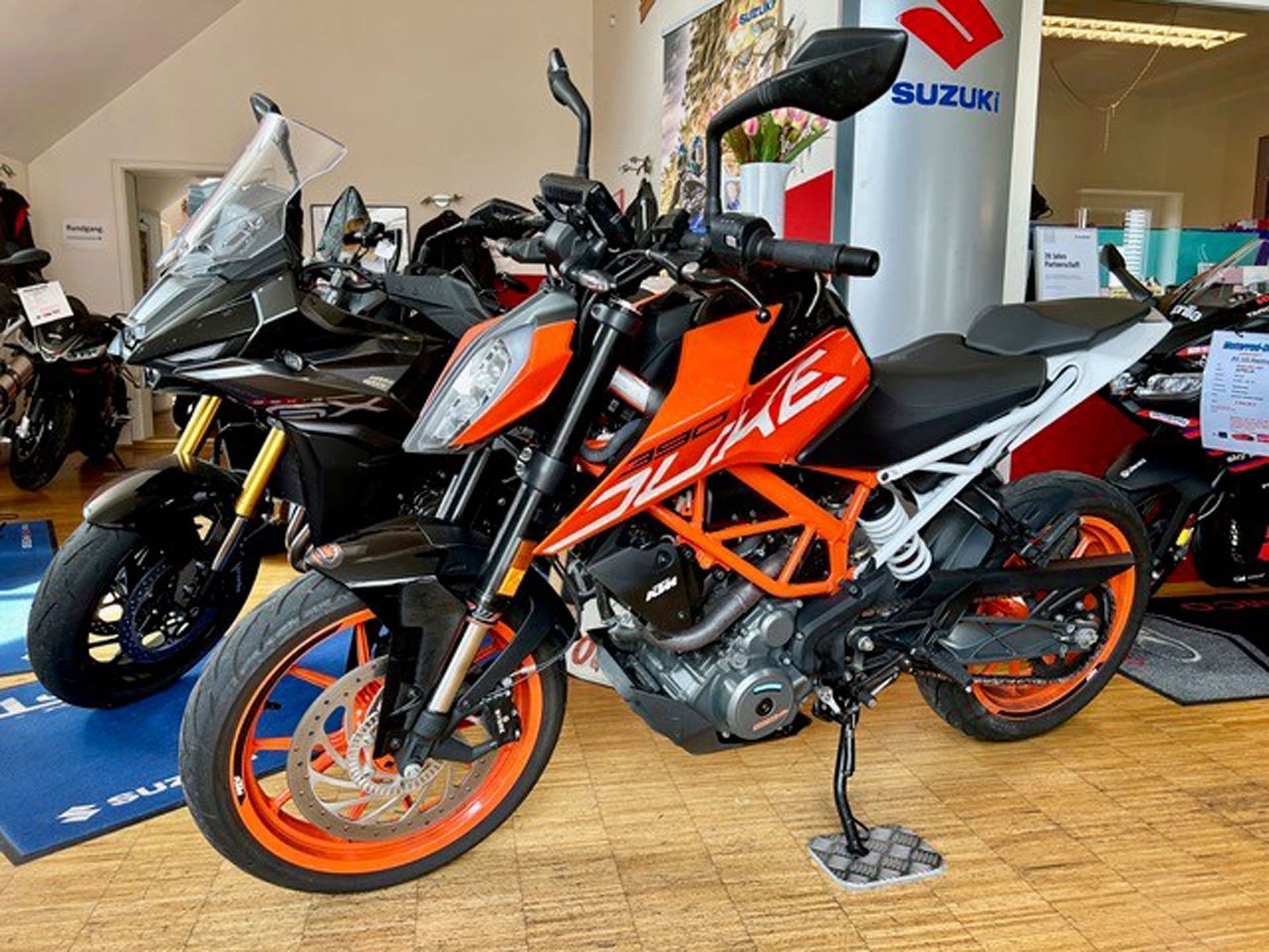 Orange-schwarzes KTM 390 Duke-Motorrad mit einem schwarz-goldenen Adventure-Motorrad im Hintergrund, in einem Autohaus.