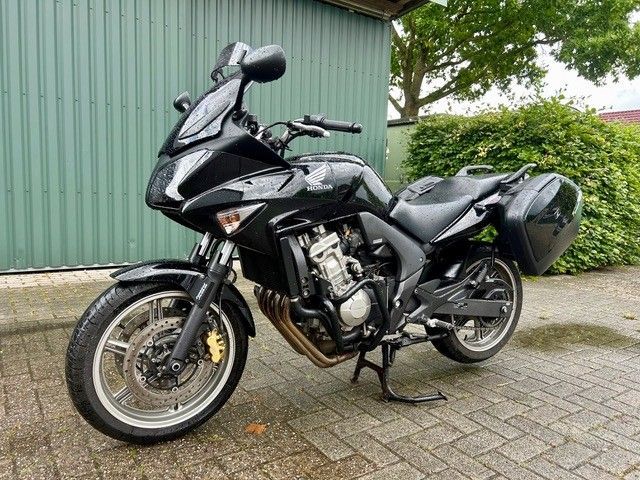 Schwarzes Tourenmotorrad parkt auf einer gepflasterten Auffahrt neben einer grünen Wellblechmauer.