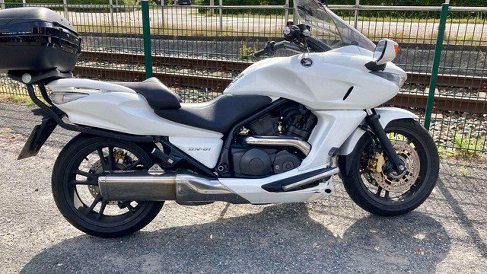 Weißes Honda-Motorrad, neben Bahngleisen geparkt, mit einer schwarzen Aufbewahrungsbox auf der Rückseite.