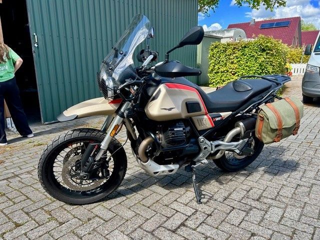 Ein Adventure-Motorrad mit Seitentaschen und hoher Windschutzscheibe parkt auf einem Backsteinpflaster neben einem grünen Gebäude.