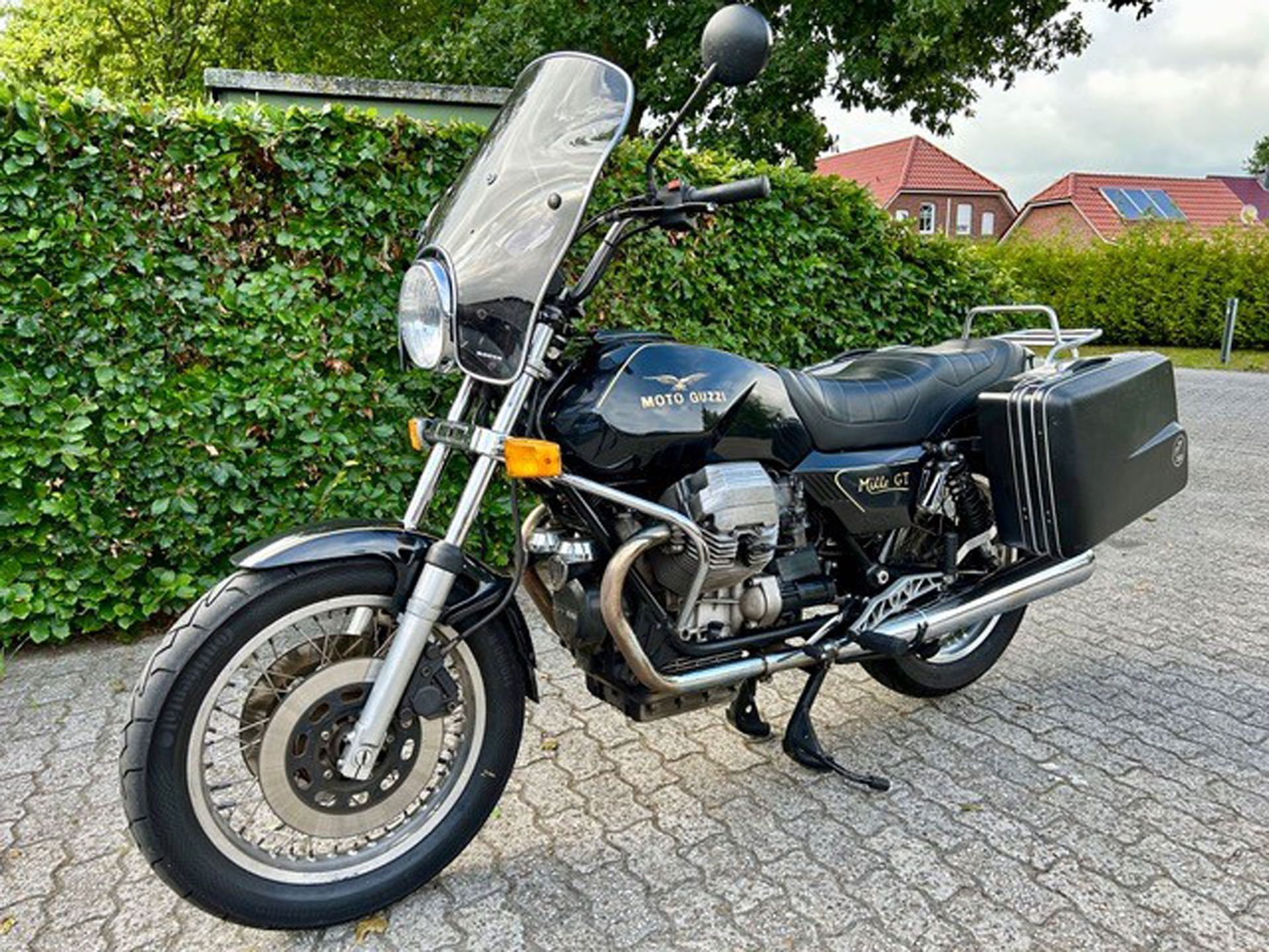 Schwarzes Moto Guzzi-Motorrad, geparkt auf einer Ziegelfläche, mit Windschutzscheibe, Satteltaschen und Chrom-Akzenten.