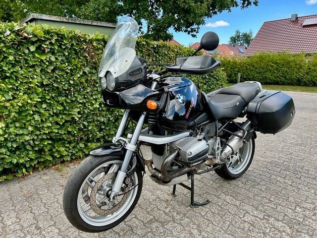Schwarzes Motorrad BMW R1100R auf Ziegelpflaster geparkt, im Hintergrund eine grüne Hecke.