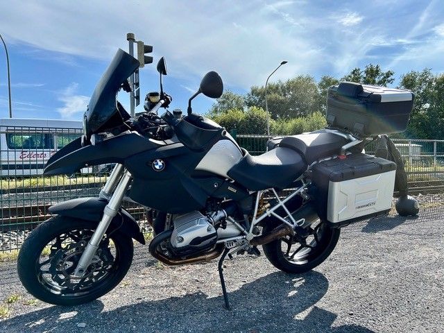 Schwarzes BMW-Motorrad mit Gepäck, das im Freien in der Nähe eines Zauns und einer Eisenbahnstrecke unter blauem Himmel geparkt ist.