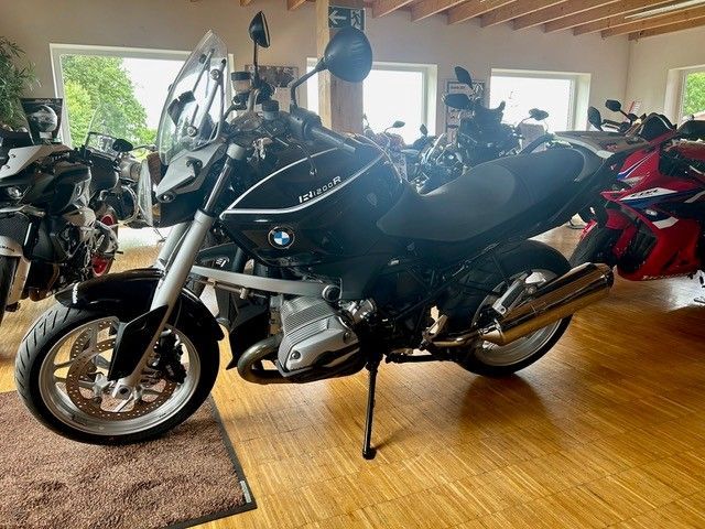 Schwarzes Motorrad in einem Ausstellungsraum, im Hintergrund weitere Motorräder.