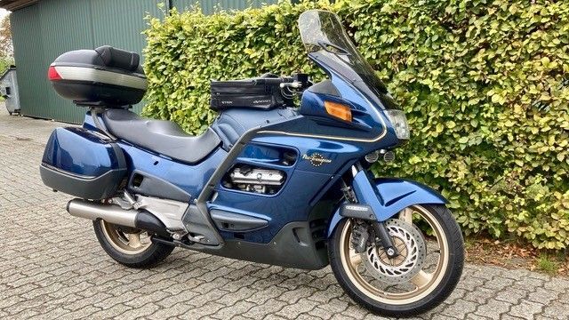 Blaues Honda ST1100 Tourenmotorrad mit Gepäck, geparkt auf einer Ziegelfläche vor einer Hecke.