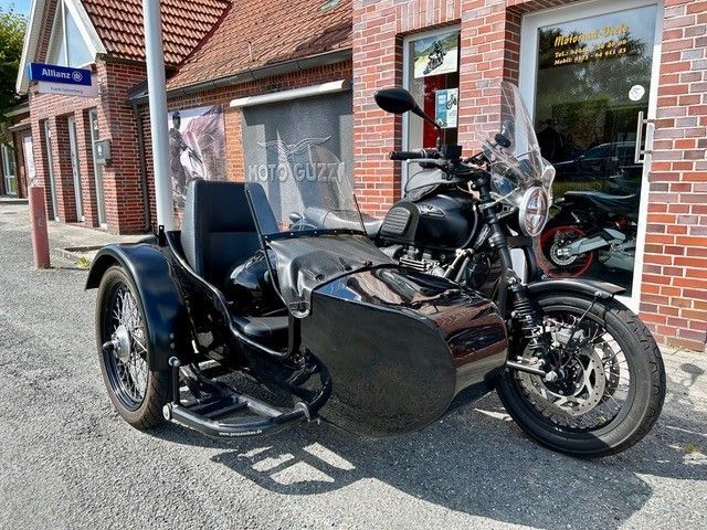 Schwarzes Motorrad mit Beiwagen, das vor einem Backsteingebäude mit dem Schild „Moto Guzzi“ geparkt ist.