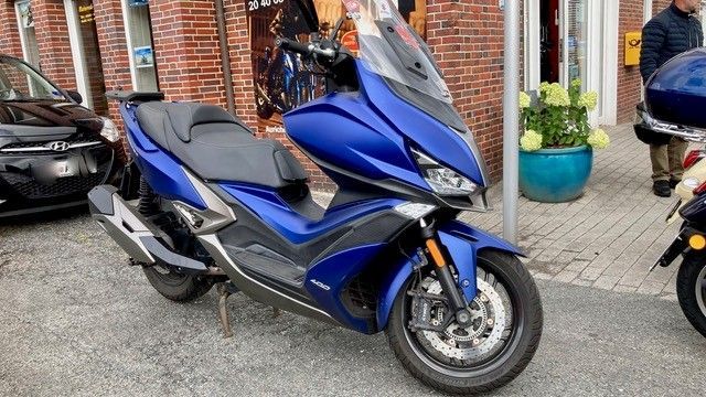 Ein blauer Tourenroller mit Windschutzscheibe und schwarzem Sitz steht auf der Straße vor einem Geschäft.