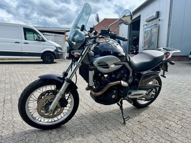 Ein schwarzes Motorrad parkt auf dem Kopfsteinpflaster neben einem weißen Lieferwagen und Gebäuden unter einem bewölkten Himmel.
