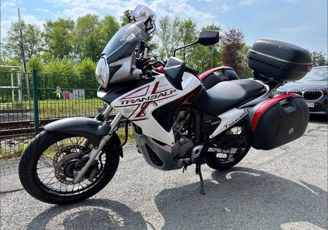 Weiß-schwarzes Honda Transalp-Motorrad mit Satteltaschen und Topcase, auf Asphalt geparkt.