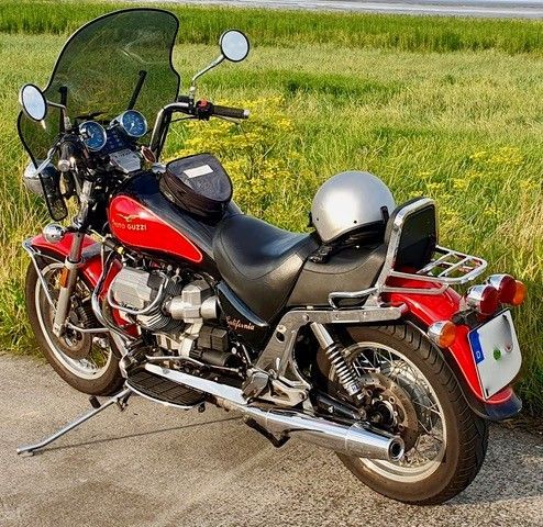 Ein rot-schwarzes Moto Guzzi-Motorrad mit schwarzer Windschutzscheibe und silbernem Helm parkt am Straßenrand.