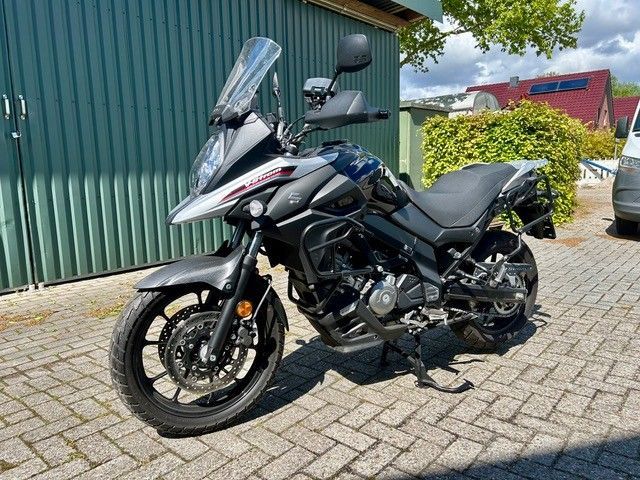 Ein schwarzes Sporttouring-Motorrad parkt auf einer gepflasterten Auffahrt neben einem grünen Schuppen und einer Hecke.