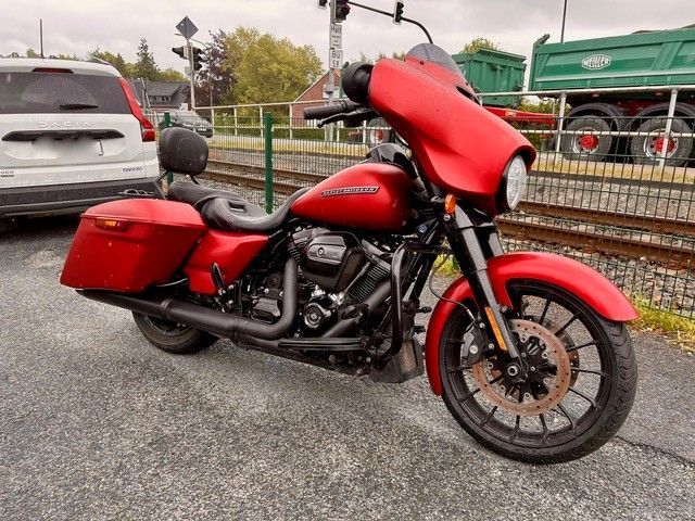 Ein rotes Tourenmotorrad parkt auf nassem Asphalt neben einem Zaun; im Hintergrund sind ein weißes Auto und ein grüner Lkw zu sehen.