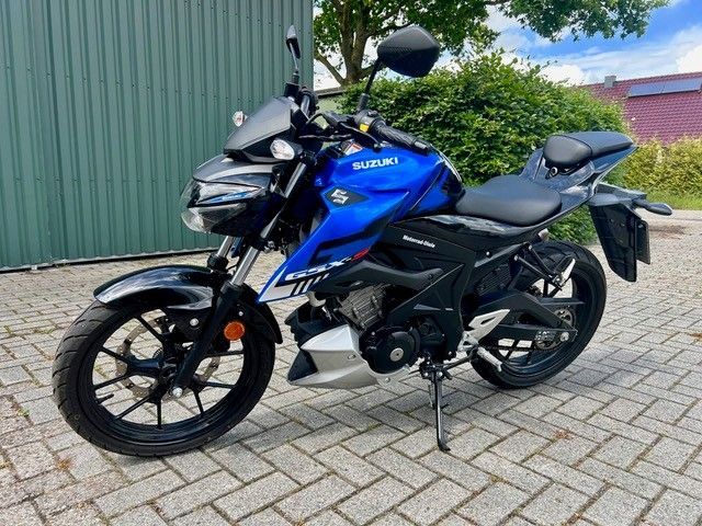 Ein blaues Suzuki-Motorrad parkt auf einer gepflasterten Auffahrt neben einer grünen Metallwand.