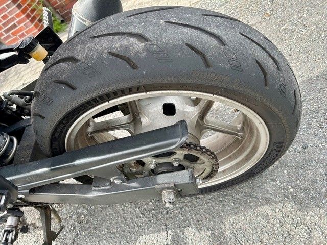 Nahaufnahme eines Motorradhinterrads, Reifens und Schwingarms auf dem Asphalt in der Nähe einer Backsteinmauer.