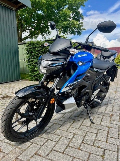 Ein blauer und schwarzer Roller parkte auf einer gepflasterten Auffahrt neben einem grünen Gebäude und Bäumen.