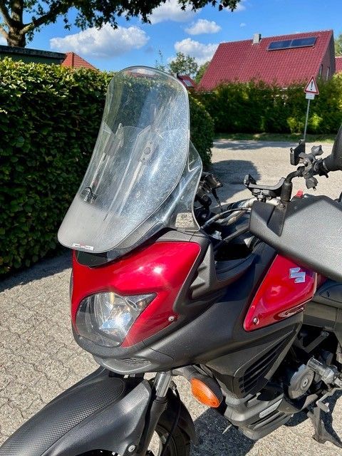 Ein roter und schwarzer Roller mit hoher Windschutzscheibe parkt auf einer sonnigen Straße neben Hecken und Häusern.