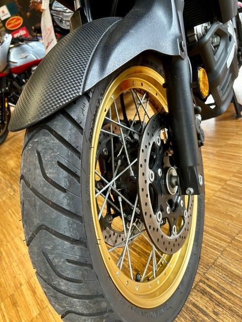 Nahaufnahme eines Motorradvorderrads mit goldener Felge und schwarzem Reifen auf einem Holzboden.