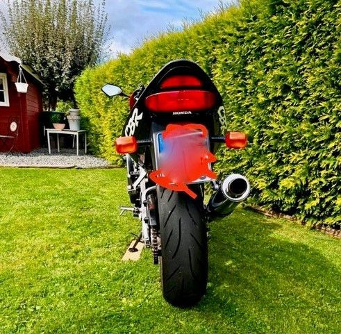 Schwarz-rotes Motorrad auf grünem Gras geparkt, vor einem roten Schuppen und grünen Büschen.
