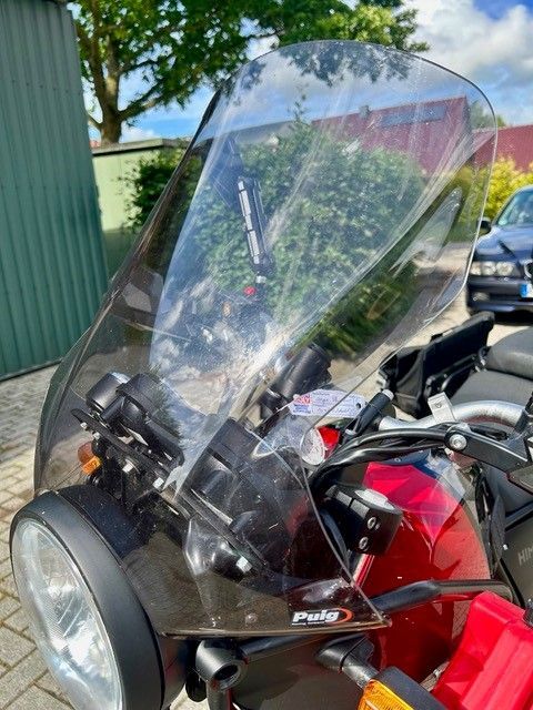Rotes Motorrad mit hoher, durchsichtiger Windschutzscheibe, im Freien an einem grünen Zaun und Baum geparkt.