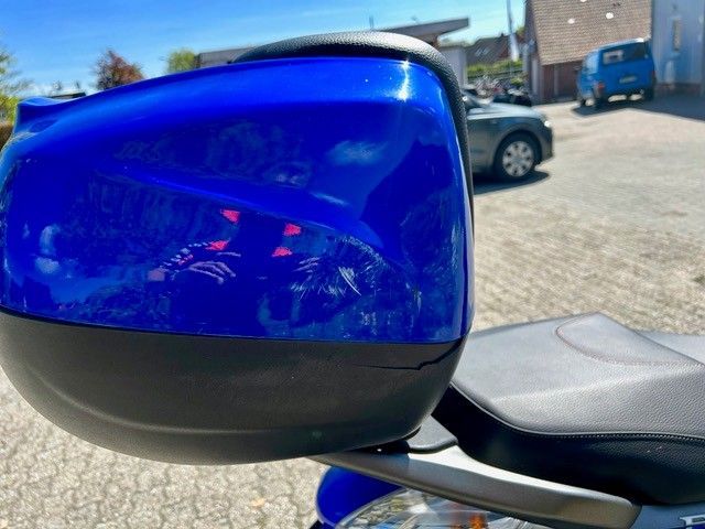 Auf einem Parkplatz steht ein blaues Motorrad, im Hintergrund ist ein Auto zu sehen.