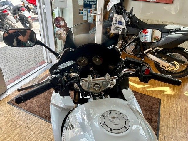 Blick aus dem Cockpit eines Motorrads auf einen weißen Kraftstofftank und Lenker in einer Garage, in deren Nähe sich weitere Motorräder befinden.