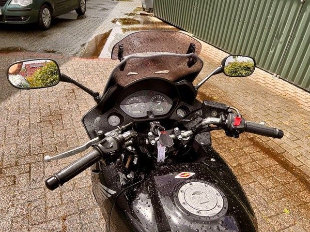 Motorradcockpit im Regen, von oben betrachtet; geparkt auf einem nassen Backsteinweg neben einem grünen Zaun.
