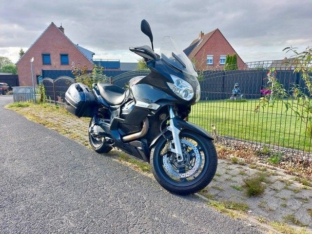Schwarzes Tourenmotorrad auf Asphalt neben einem grünen Hof geparkt, im Hintergrund Häuser.