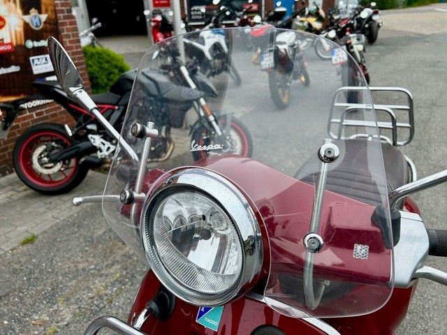 Ein roter Motorroller mit durchsichtiger Windschutzscheibe ist vor einer Reihe Motorräder geparkt.