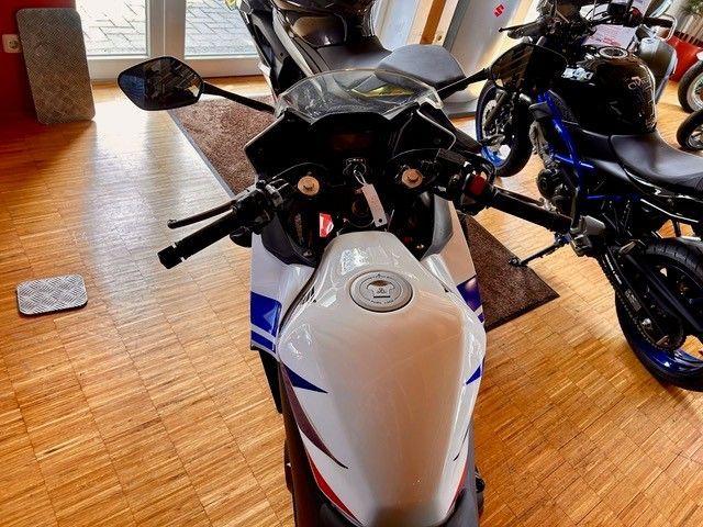 Ein weiß-blaues Sportmotorrad, von oben betrachtet, in einem Ausstellungsraum, in dessen Nähe sich weitere Motorräder befinden.