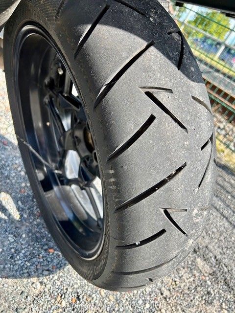 Nahaufnahme eines Motorradreifens mit Profilrillen, auf einer Asphaltoberfläche, abgewinkelte Ansicht, sonniger Tag.