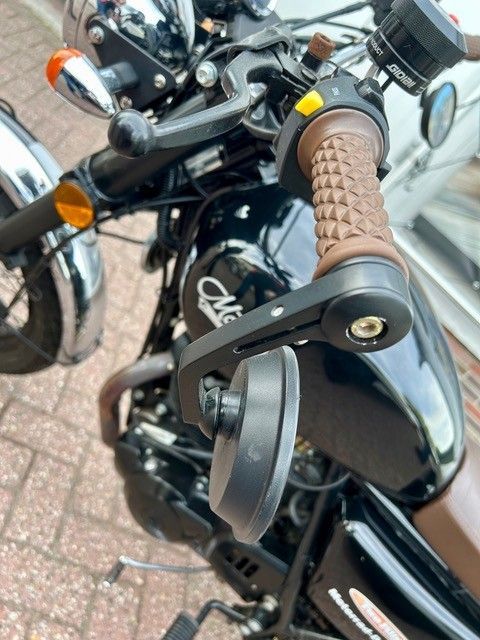 Nahaufnahme eines schwarzen Motorrads mit braunen Griffen und Spiegel, das auf einem gepflasterten Gehweg geparkt ist.