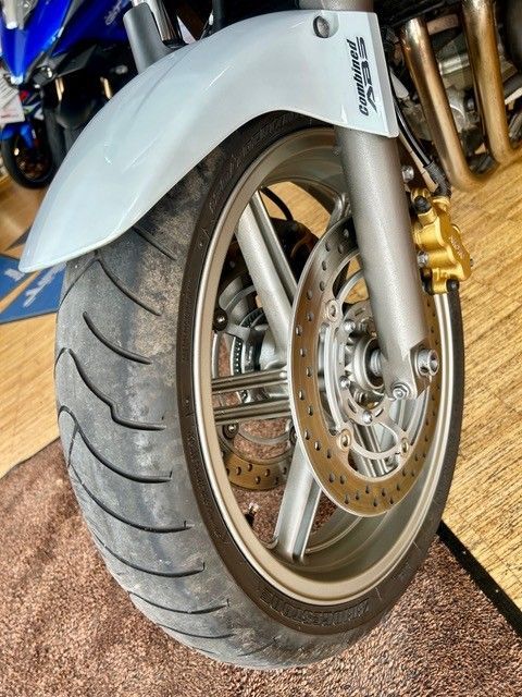 Nahaufnahme eines Motorradvorderrads mit silbernem Schutzblech und goldenem Bremssattel in einem Innenraum