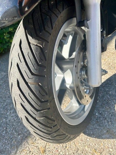 Nahaufnahme eines Motorradvorderrads mit Reifen auf Asphalt, Felge und Bremsscheibe sind zu sehen.