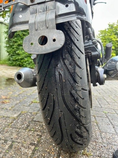 Nahaufnahme eines Motorradhinterreifens auf einer gepflasterten Auffahrt, die das Profil und das Auspuffrohr zeigt.