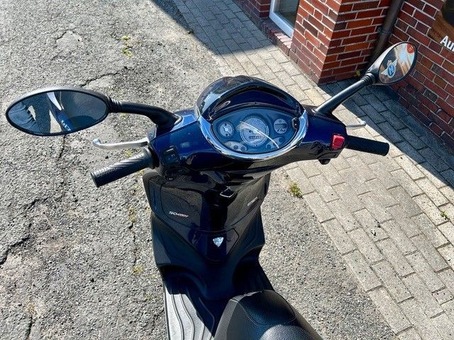 Ein blauer Motorroller ist auf einem gepflasterten Gehweg vor einem Backsteingebäude geparkt.