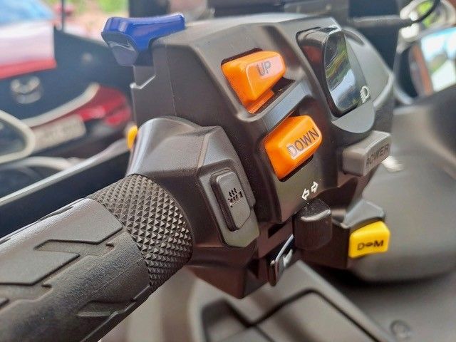 Nahaufnahme der Lenkersteuerung eines Motorrads mit orangefarbenen und blauen Tasten, einem strukturierten Griff und einem schwarzen Gehäuse.