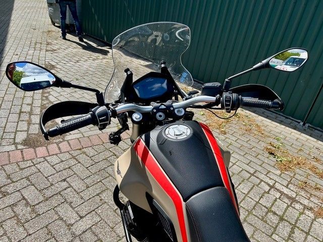 Motorradcockpit mit Spiegeln und Windschutzscheibe, geparkt auf einer gepflasterten Straße neben einem grünen Zaun