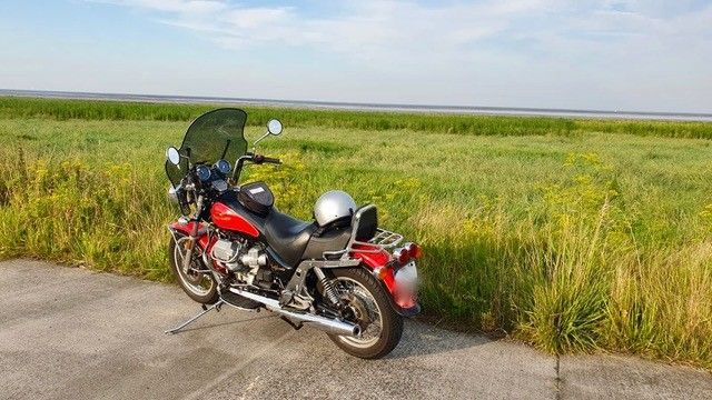Ein rotes Motorrad parkt am Straßenrand neben einem Feld, der Helm liegt auf dem Sitz, unter blauem Himmel.