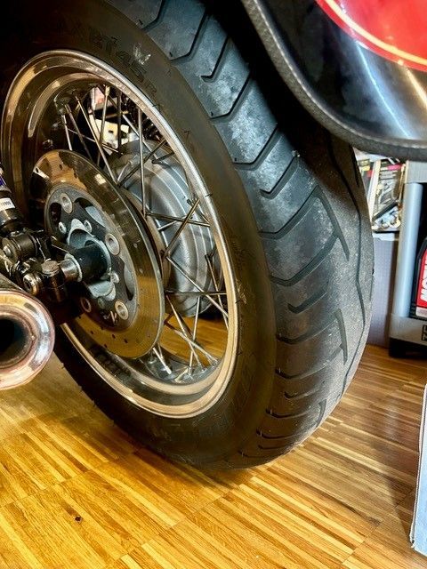 Nahaufnahme eines Motorradhinterrades mit verchromten Speichen und Reifen auf einem Holzboden.