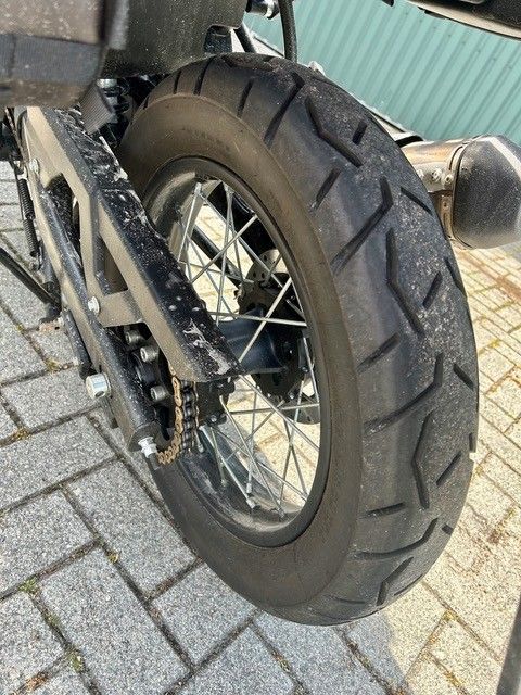Nahaufnahme eines Motorradhinterrads mit Reifen auf asphaltierter Fläche, Kette und Speichen sind sichtbar.