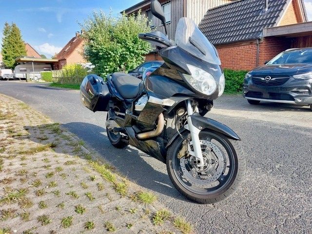 Schwarzes Moto Guzzi-Tourenmotorrad, mit Satteltaschen, Windschutzscheibe und Wohnhaus im Hintergrund auf einer Straße geparkt.