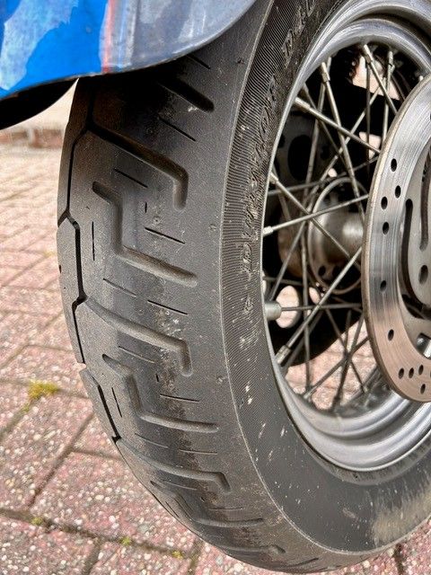 Nahaufnahme eines Motorradreifens mit schwarzer Seitenwand und Profilmuster. Der Reifen ist auf einem Speichenrad mit einer silbernen Bremsscheibe montiert.