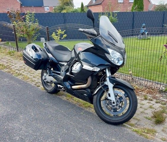 Schwarzes Moto Guzzi-Motorrad, geparkt auf Asphalt neben einem grasbewachsenen Hof, mit einem Zaun und Häusern im Hintergrund.