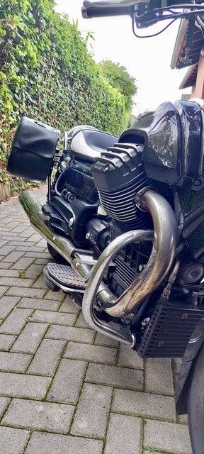 Schwarzes Motorrad, geparkt auf einem gepflasterten Platz neben einer grünen Hecke.